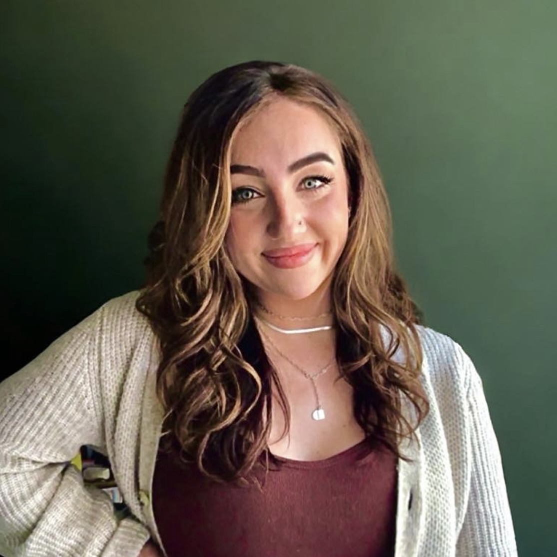 A woman with brown hair and a burgundy shirt with a tan sweater poses softly in front of a green backdrop.s